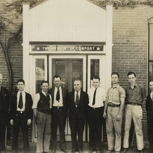 Group of white men in suits and ties standing in line outside brick building with glass door