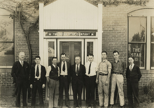 Group of white men in suits and ties standing in line outside brick building with glass door