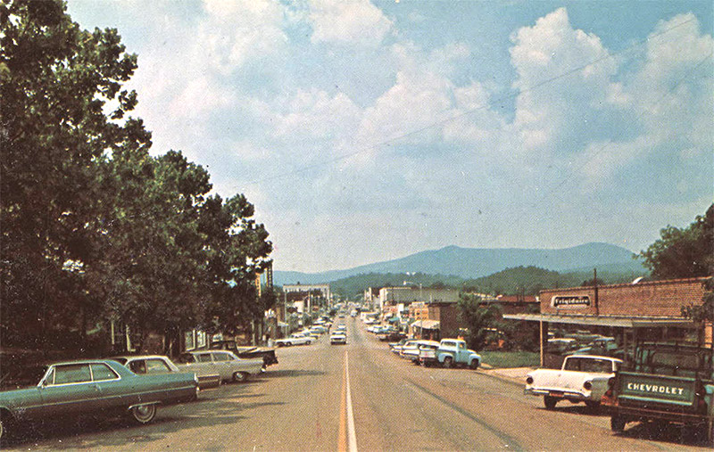 Cars parked outside town buildings on two-lane street with hills in the background