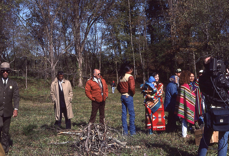Native American women in traditional clothing and men at mound site with white camera crew and African-American park ranger