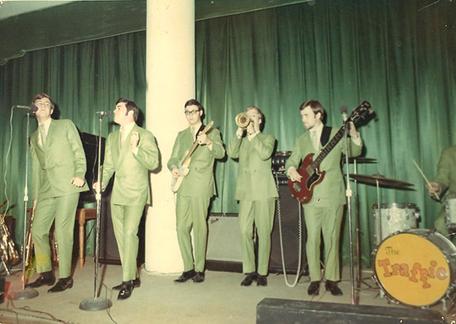 Five young men in green suits playing instruments and singing on stage in front of a green curtain