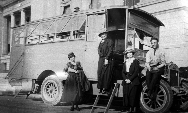 White man and three white women in hats and dresses posing with bus