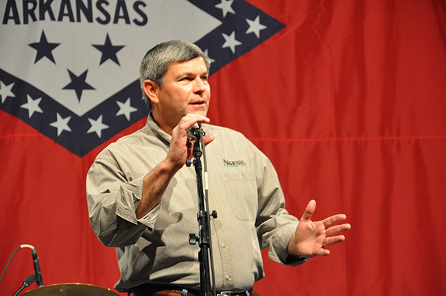 White man speaking at microphone with Arkansas flag background behind him