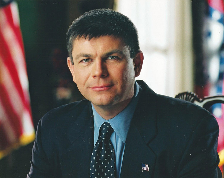 White man in suit and tie with flag behind him