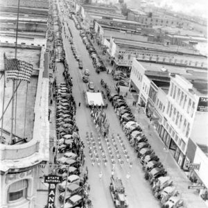 Aerial view of parade proceeding along a road lined with buildings and parked cars