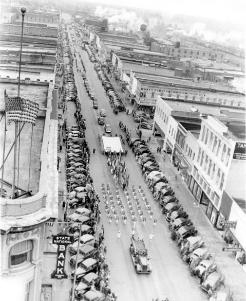 Aerial view of parade proceeding along a road lined with buildings and parked cars