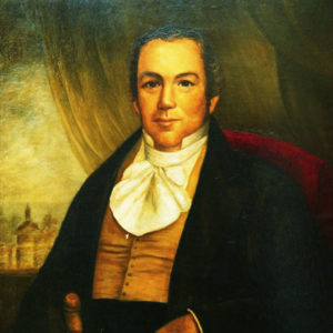 White man in black suit with a white cravat and curtains in the background