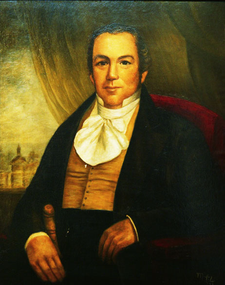 White man in black suit with a white cravat and curtains in the background
