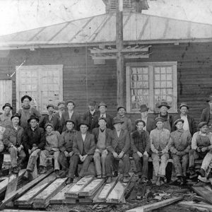 White men sitting before single story wooden building with wooden planks lying about