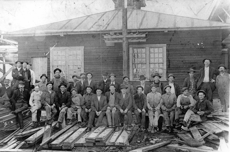 White men sitting before single story wooden building with wooden planks lying about