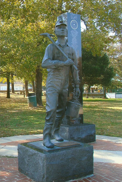 Statue of man with mining equipment on base and polished stone column behind it