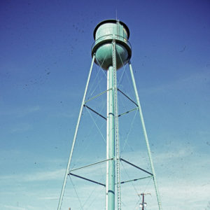 Green water tower and brick building inside fence with person standing by fence