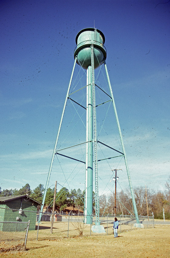 Green water tower and brick building inside fence with person standing by fence
