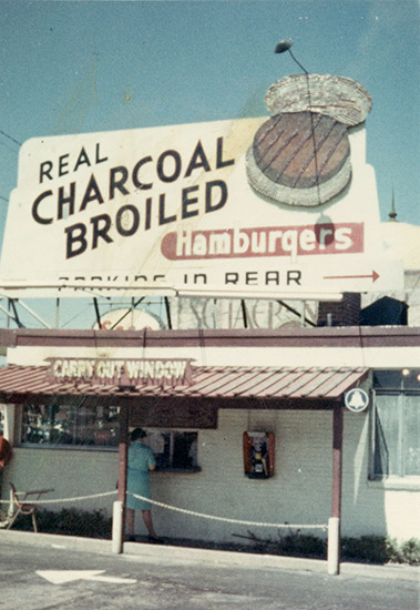 Small brick building with awning with "Real charcoal broiled hamburgers" and "carry out window" signs above it