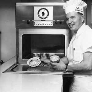 White man in chef's uniform with chef's hat saying "Lido" putting food in cardboard cups in an oven