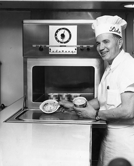White man in chef's uniform with chef's hat saying "Lido" putting food in cardboard cups in an oven