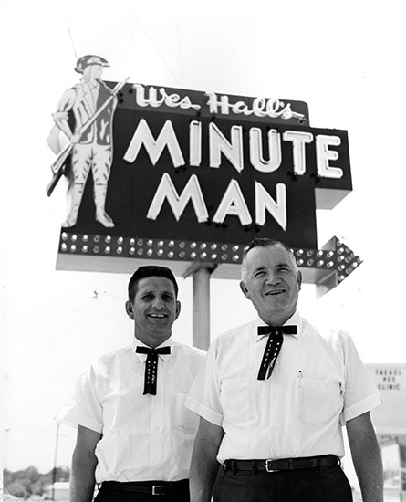 Young white man and older white man in matching white shirt and black pants uniforms with ties under "Wes Hall's Minute Man" sign