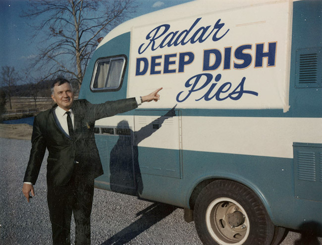 White man in suit and tie pointing at "Radar deep dish pies" advertisement on a recreational vehicle