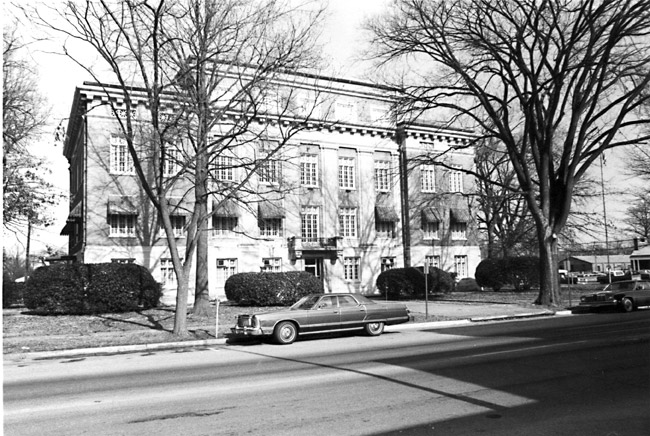 Multistory brick building with half sized upper story first floor window awnings and bare trees and cars parked in front