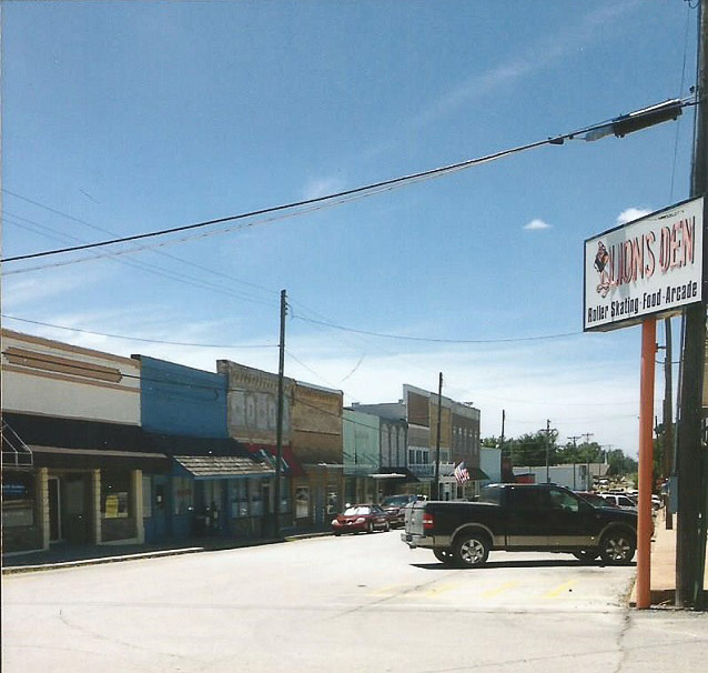 Storefronts on street with parked cars and blue skies and sign reading "Lions Den Roller Skating Food Arcade"