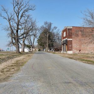 gravel road with grass field and trees with red brick building and barn on right