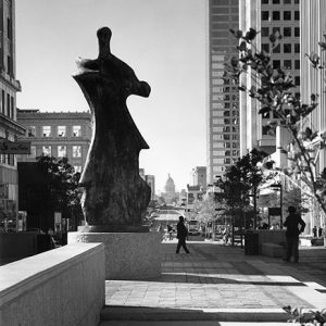 Sculpture on pedestal surrounded by tall buildings with state capitol building in background