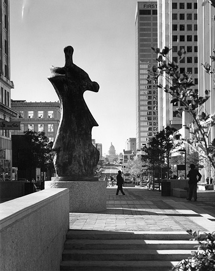 Sculpture on pedestal surrounded by tall buildings with state capitol building in background