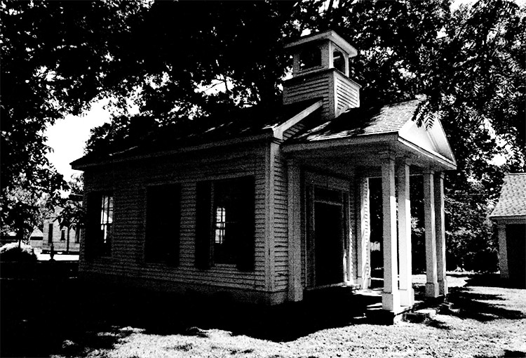 Single-story chapel building with cupola and covered porch next to house and tree