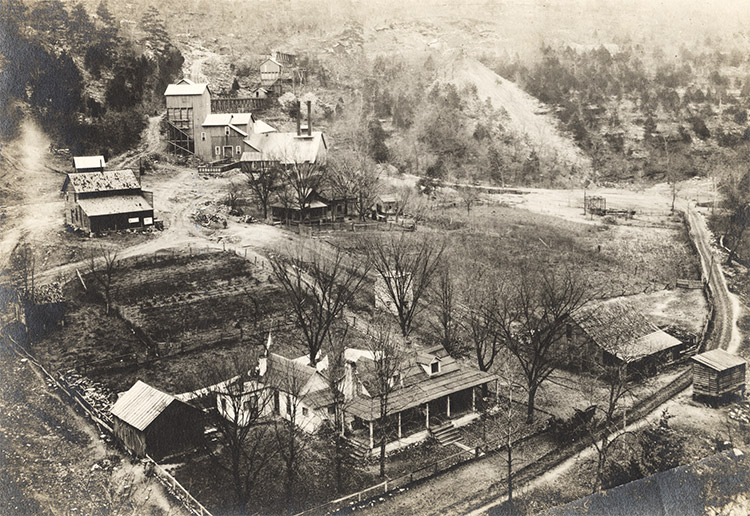 Aerial view of mining buildings and multistory house with covered porch outbuildings and garden