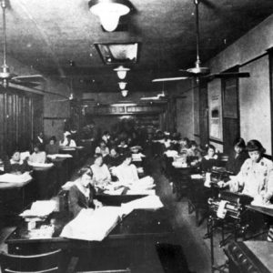Three rows of African-American women working at their desks in long hall with lights and ceiling fans