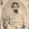 White man with mustache and beard in gray military uniform holding sword