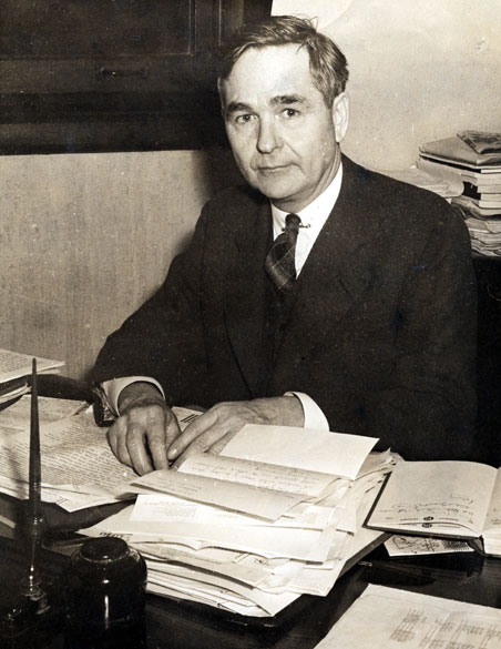 White man in suit and tie with desk covered in papers and open books