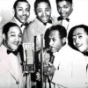 African-American man playing saxophone with five African-American men in suits and bow ties in front of a CBS microphone