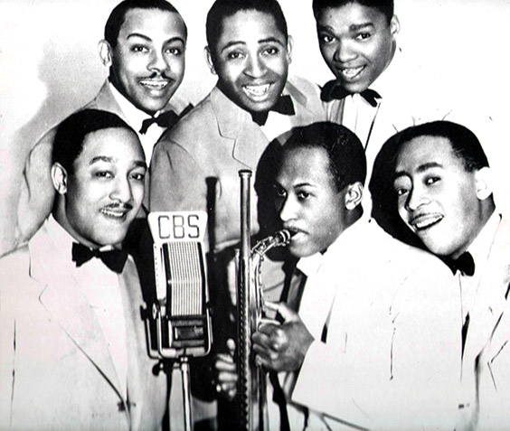 African-American man playing saxophone with five African-American men in suits and bow ties in front of a CBS microphone