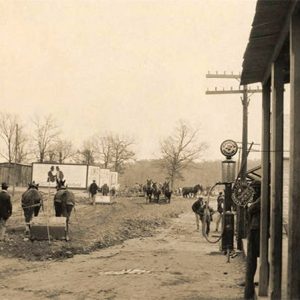 Men and horses on dirt roads with house and barn in the background and storefront in the foreground