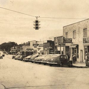 Traffic light hanging over town street with parked cars outside brick storefronts and people gathered on street corner in the foreground