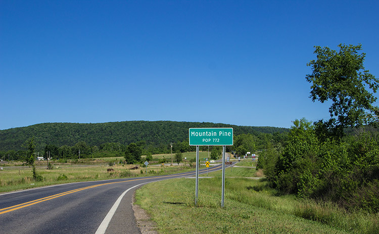 Two-lane highway through countryside with green "Mountain Pine" sign on the right and tree covered hills in the distance