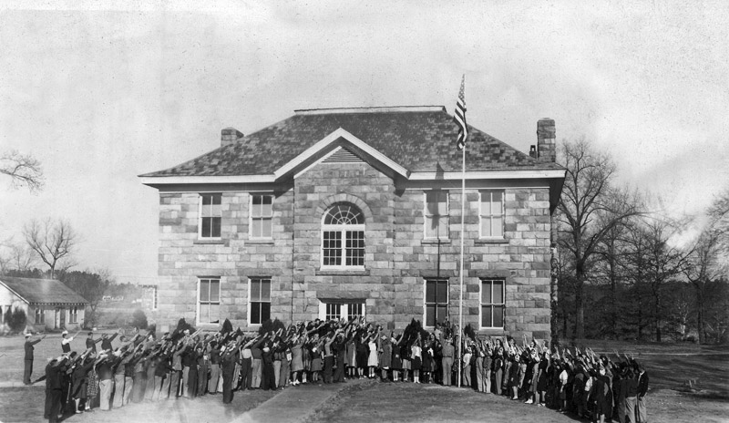 large group of white teenagers outside two-story stone building doing old-fashioned salute of American flag on flagpole, with right arms extended straight out