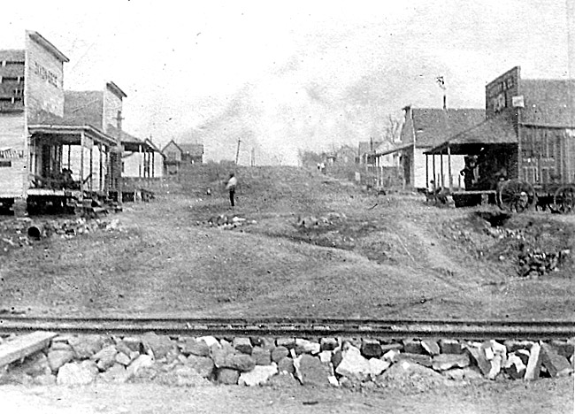 Dirt uphill road with buildings on both sides and man standing and railroad tracks