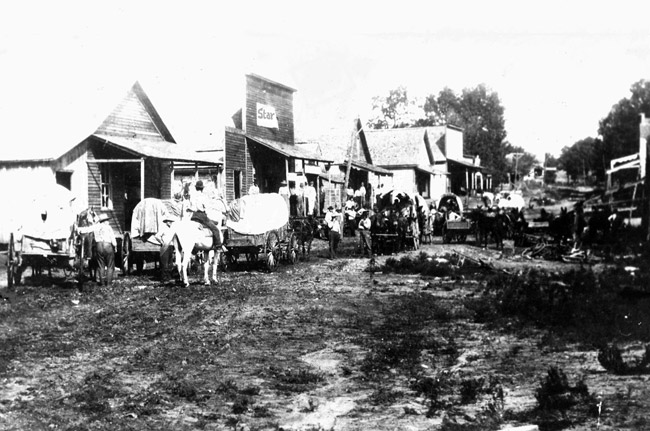 Crowded dirt road with storefront buildings horses and covered wagons