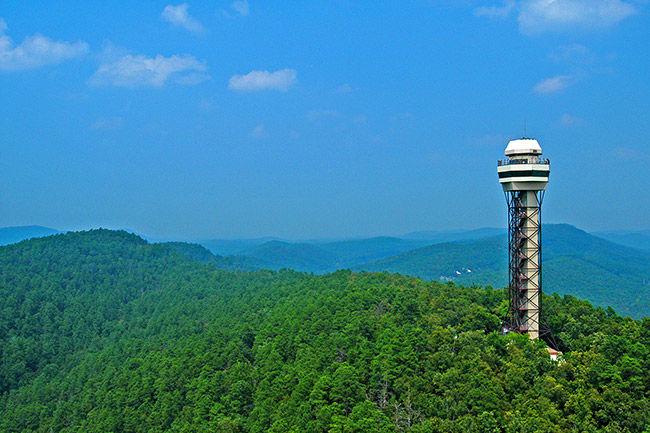 Observation tower on mountain top with tree covered mountains in the distance and blue skies