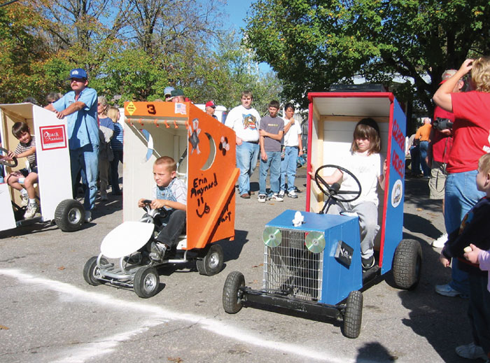White children on go-carts modified to look like "outhouses" sitting at the starting line with crowd behind them