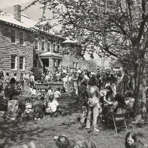 Crowd of people sitting and standing under trees outside multistory stone building with covered porch