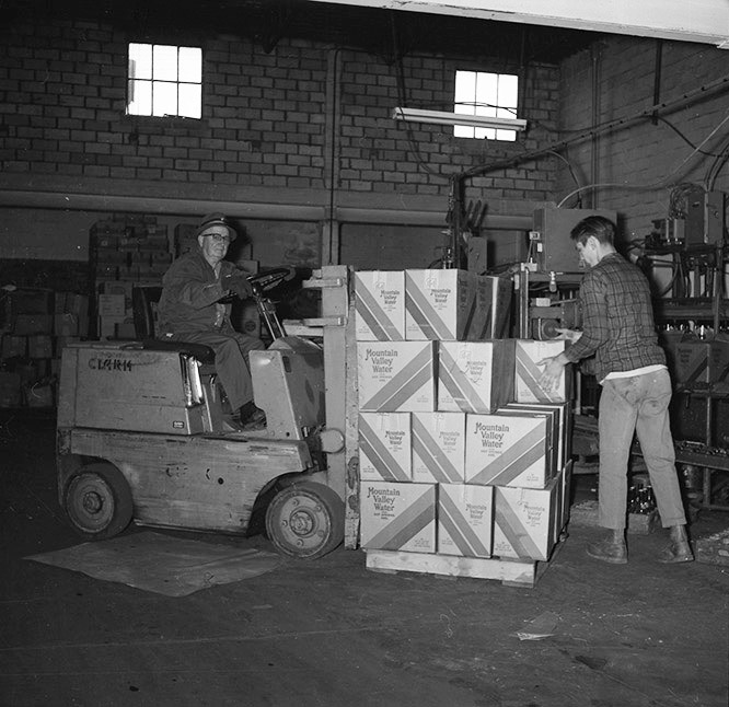 Old white man with glasses and hat moving boxes with forklift in bottling plant