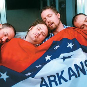Four white men sleeping under Arkansas flag blanket