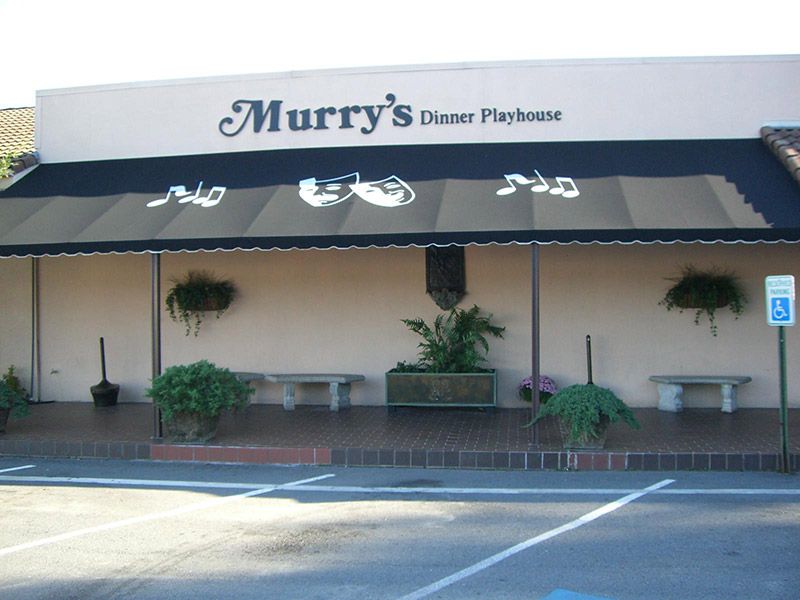 Exterior of "Murry's dinner playhouse" building with decorative plants and benches under its awning