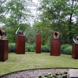 Five sculptures resembling tree sections with stands of various dimensions in landscaped sculpture garden by forest