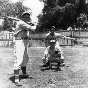 Young white catcher umpire and batter in uniforms playing baseball with wooden fence behind them