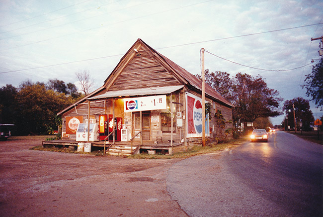 Single-story storefront with open gable roof and covered porch and prominent Pepsi signs on rural road