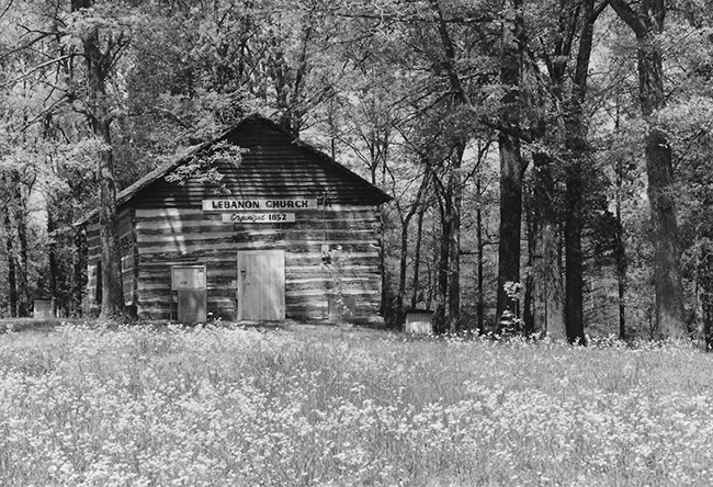 Single-story cabin building with "Lebanon Church organized 1852" sign under trees in field